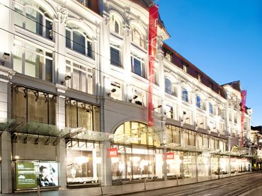 Beleuchtete Fassade eines großen, eleganten Einkaufszentrums am Abend, mit gewölbten Fenstern und Glasdächern. Die Straße ist leer, und die Schaufenster sind hell beleuchtet.