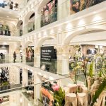 Ein elegantes mehrstöckiges Einkaufszentrum mit klassischen architektonischen Details. Käufer können in Geschäften mit großen Glasfenstern nach Kleidung stöbern. Um das offene Atrium herum sind dekorative Pflanzen und Blumen aufgestellt.