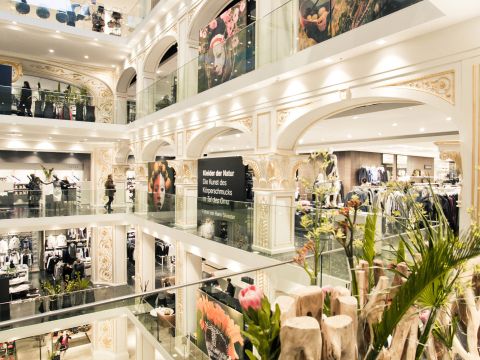 Ein elegantes mehrstöckiges Einkaufszentrum mit klassischen architektonischen Details. Käufer können in Geschäften mit großen Glasfenstern nach Kleidung stöbern. Um das offene Atrium herum sind dekorative Pflanzen und Blumen aufgestellt.