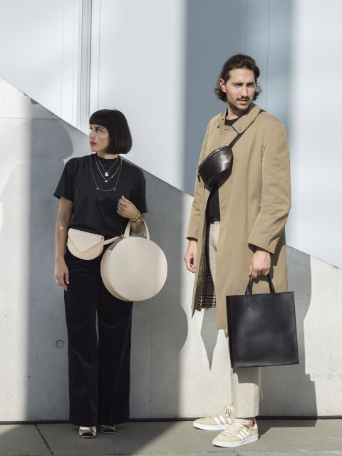 Ein modisch gekleideter Mann und eine Frau stehen im Freien. Die Frau trägt ein schwarzes Oberteil und hält eine beige runde Tasche. Der Mann trägt einen beigen Mantel, eine schwarze Tragetasche und eine Umhängetasche. Beide blicken in die gleiche Richtung und stehen vor einer weißen Wand.