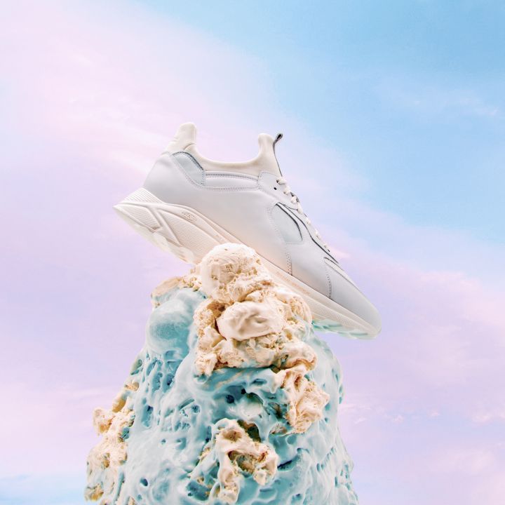 Ein weißer Turnschuh wird auf kreative Weise auf einem Hügel präsentiert, der an geschmolzenes Eis erinnert, vor einem himmelblauen und pastellrosa Hintergrund, was eine ätherische und skurrile Atmosphäre vermittelt.