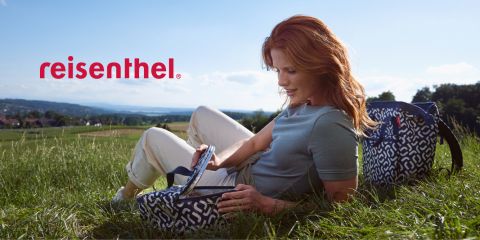 Eine Person mit langen roten Haaren, die ein hellblaues Hemd und weiße Hosen trägt, entspannt sich im Gras und hält eine gemusterte Tasche in der Hand. Sie steht draußen auf einem sonnigen Feld mit Blick auf die Hügel in der Ferne. Das Wort „Reisenthel“ ist in roter Schrift zu sehen.