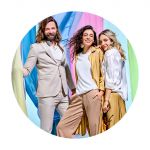 Drei Personen posieren in stylischen, hellen Outfits vor einem farbenfrohen Hintergrund aus drapierten Stoffen in Pastelltönen. Die Szene vermittelt eine fröhliche und modische Atmosphäre mit Fokus auf zeitgenössischem Stil.