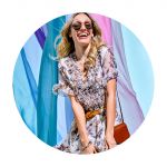 Eine lächelnde Frau mit Sonnenbrille und geblümtem Kleid steht vor einem Hintergrund aus farbenfrohem, fließendem Stoff. Sie trägt eine braune Umhängetasche und ihr blondes Haar wird sanft vom Wind weht.