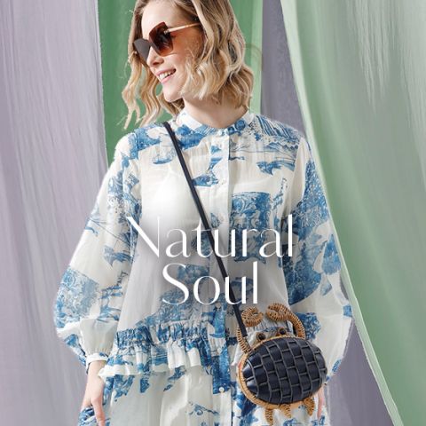 Eine Person mit Sonnenbrille trägt ein weiß-blau gemustertes Kleid und steht vor einem Hintergrund aus fließendem grün-violettem Stoff. Sie trägt eine kleine, runde gewebte Handtasche. Der Text auf dem Bild lautet „Natural Soul“.