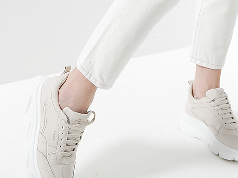Eine Person in weißen Turnschuhen und weißer Hose steht auf einer weißen Fläche. Die Turnschuhe haben eine dicke Sohle und Schnürsenkel und ihr Design ist minimalistisch. Das Bild konzentriert sich auf das Schuhwerk und die Unterschenkel.