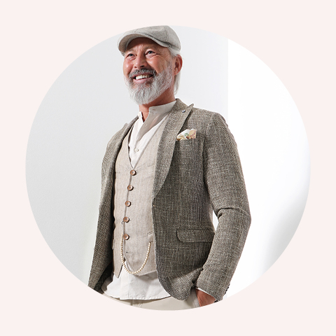 Älterer Mann in Tweedjacke, beiger Weste und Schiebermütze, der selbstbewusst lächelt. Der Hintergrund ist in sanftem Weiß gehalten und vermittelt ein modernes und elegantes Gefühl.