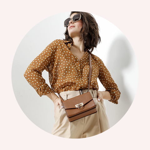 Eine Frau mit Sonnenbrille und einer braunen Bluse mit Tupfenmuster, kombiniert mit beigen Hosen, posiert selbstbewusst mit einer Umhängetasche aus braunem Leder. Der Hintergrund ist ein einfacher Lichtkreis.