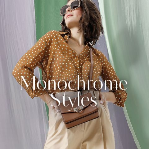 Eine Person mit Sonnenbrille, braunem Polka Dot-Shirt und beiger Hose posiert selbstbewusst mit den Händen auf den Hüften. Eine braune Umhängetasche ist zu sehen. Der Hintergrund ist mit fließenden grünen und grauen Stoffen gestaltet. Der Text lautet „Monochrome Styles“.