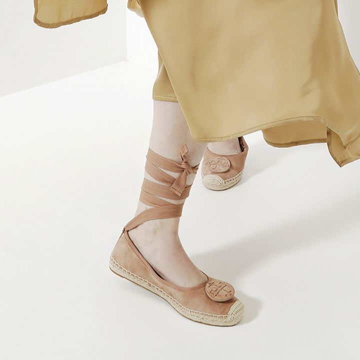 Nahaufnahme einer Person, die hellbraune Espadrilles mit um den Knöchel gewickelten Schnürsenkeln trägt. Die Schuhe haben auf der Vorderseite dekorative Muster. Die Person steht auf einem weißen Boden und trägt einen fließenden hellbraunen Rock.