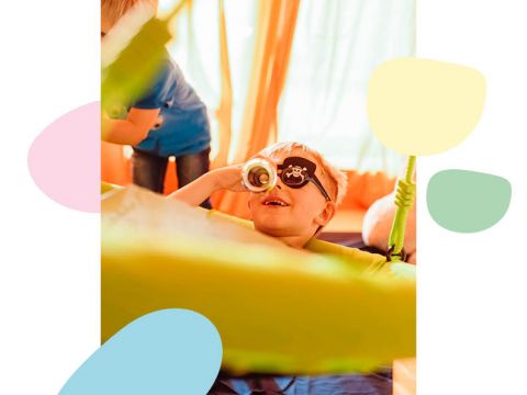 Ein Kind mit einer Piratenaugenklappe und einem Teleskop in der Hand schaut spielerisch hindurch. Es ist von bunten Formen und sanftem Licht umgeben, im Hintergrund ist teilweise ein weiteres Kind zu sehen.