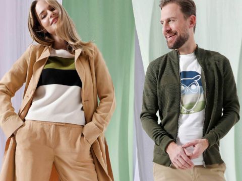 Eine Frau in hellbraunem Mantel und hellbrauner Hose sowie einem weißen Pullover posiert mit einer Hand auf der Hüfte. Ein Mann in grüner Jacke und heller Hose lächelt zur Seite. Sie stehen vor einem Hintergrund aus grün und weiß drapiertem Stoff.