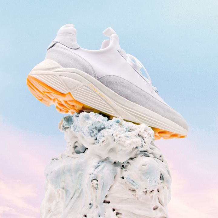 Ein weiß-grauer Turnschuh mit orangefarbenen Sohlen steht auf einem Hügel aus strukturiertem weißem Material, das an Eiscreme erinnert. Der Hintergrund ist ein pastellfarbener Himmel mit zarten Rosa- und Blautönen.