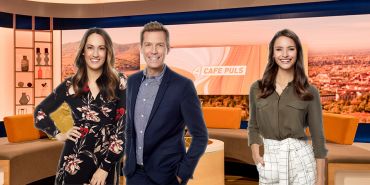 Drei Personen stehen und lächeln in einem Fernsehstudio, im Hintergrund läuft auf einem Bildschirm „Cafe Puls“. Das Studio ist warm beleuchtet und minimalistisch eingerichtet. Die Personen sind stilvoll und leger gekleidet.