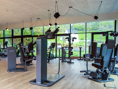 Ein modernes Fitnessstudio mit großen Fenstern, die einen malerischen Blick nach draußen bieten. Das Fitnessstudio ist mit verschiedenen Trainingsgeräten ausgestattet, darunter Laufbänder, Heimtrainer und Gewichthebergeräte. Der Raum ist mit natürlichem Licht durchflutet.