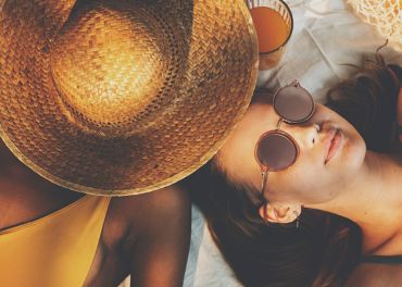 Zwei Menschen entspannen auf einer Stranddecke. Einer trägt einen großen Strohhut, der sein Gesicht verdeckt, der andere trägt eine Sonnenbrille und liegt daneben. Beide tragen Badebekleidung und haben ein Glas Orangensaft in der Nähe, was für eine sommerliche und entspannte Stimmung sorgt.