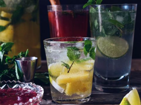 Eine Auswahl farbenfroher Cocktails auf einem Tisch. Die Getränke sind mit Ananasstücken, Limettenscheiben und frischen Minzblättern garniert. Daneben stehen ein kleiner Metallmessbecher und eine Schale mit roter Flüssigkeit. Der Hintergrund ist schwach beleuchtet.