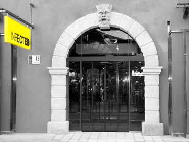 Ein Schwarzweißbild eines steinernen Torbogens mit einem stilisierten Gesicht oben drauf. Der Torbogen führt in ein Geschäft mit Glastüren. Links ist ein gelbes „INFEKTIERT“-Schild. Im Inneren sind Spiegelbilder von Menschen zu sehen.