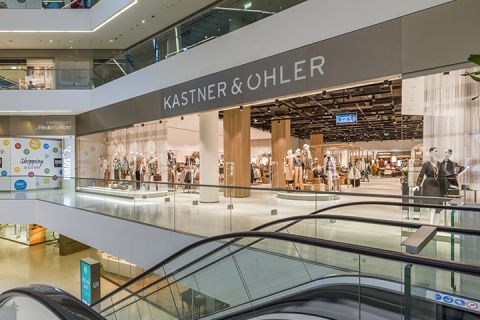 Innenansicht eines modernen Einkaufszentrums mit dem Geschäft Kastner & Öhler. An der Fassade des Ladens sind Schaufensterpuppen in verschiedenen Outfits zu sehen. Im Vordergrund sind Rolltreppen zu sehen und das Einkaufszentrum hat mehrere Ebenen mit Glasgeländern.