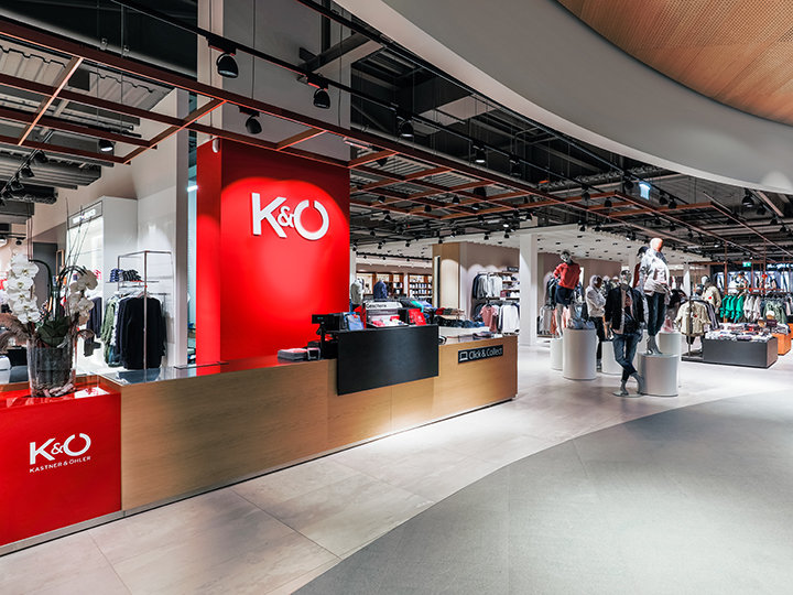 Innenansicht eines modernen Bekleidungsgeschäfts mit einer leuchtend roten Theke, auf der das K&Ö-Logo zu sehen ist. Im Laden sind mehrere Kleiderständer mit Kleidung zu sehen, im Hintergrund Schaufensterpuppen, in denen eine Person stöbert. Die Atmosphäre ist geräumig und gut beleuchtet.