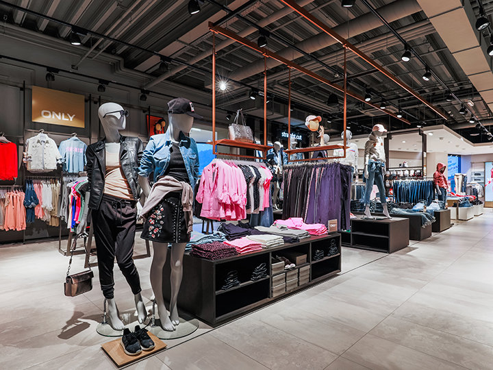 Innenansicht eines Bekleidungsgeschäfts mit Schaufensterpuppen in legerer Kleidung, darunter Jacken und Röcke. Auf Regalen und Ständern werden verschiedene Kleidungsstücke wie Hemden und Jeans ausgestellt. Das Geschäft ist modern gestaltet und verfügt über Deckenbeleuchtung und Fliesenboden.