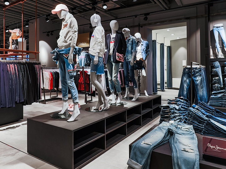 In einem Bekleidungsgeschäft sind Schaufensterpuppen in lässigen Jeans-Outfits, darunter Jeans und Jacken, zu sehen. Die umliegenden Regale enthalten verschiedene Kleidungsstücke, und auf einem Regal ist ein Stapel gefalteter Jeans zu sehen. Das Geschäft ist modern gestaltet.