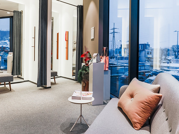 Ein moderner, stilvoller Wartebereich mit einem Sofa, einem kleinen runden Tisch und großen Fenstern, durch die man eine verschneite Stadtlandschaft sehen kann. Der Raum ist mit Teppichboden, Vorhängen und dekorativen Pflanzen auf einem Podest ausgestattet.