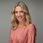 Eine Person mit langen, welligen blonden Haaren lächelt und trägt eine pfirsichfarbene Bluse. Der Hintergrund ist neutral grau.