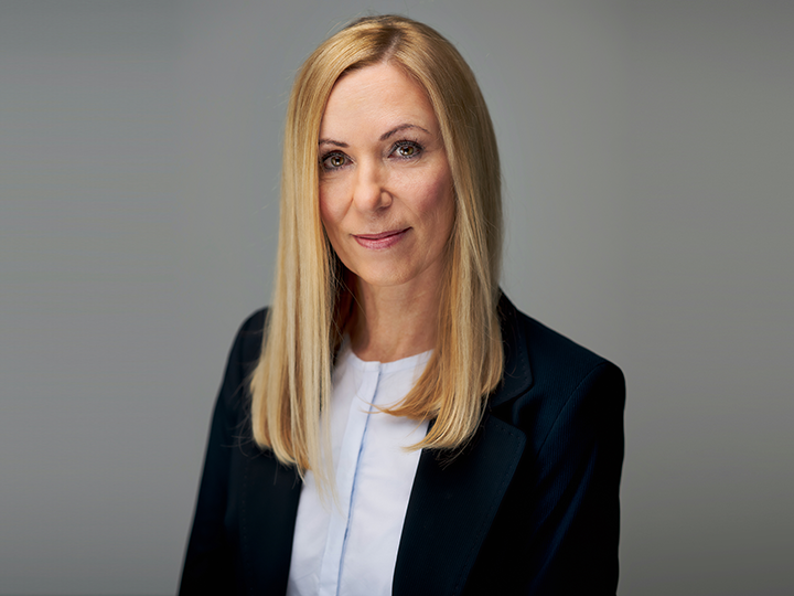 Eine Frau mit langen blonden Haaren trägt einen dunklen Blazer über einem weißen Oberteil. Sie lächelt sanft und blickt vor einem schlichten grauen Hintergrund in die Kamera.