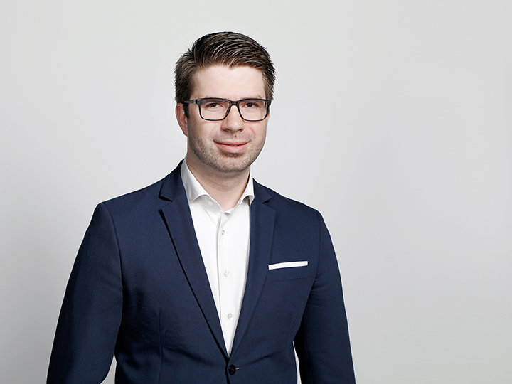 Ein Mann mit Brille, blauem Anzug und weißem Hemd steht vor einem schlichten hellen Hintergrund. Er hat kurzes braunes Haar und lächelt leicht.