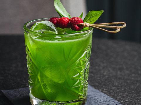 Ein leuchtend grüner Cocktail in einem strukturierten Glas, garniert mit frischen Basilikumblättern und roten Himbeeren auf einem Spieß. Er liegt auf einer dunklen Serviette vor einem verschwommenen dunklen Hintergrund.