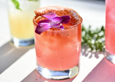 Ein leuchtend rosa Cocktail, garniert mit einer lila Blume und einem mit Gewürzen umrandeten Glas, steht auf einer sonnenbeschienenen Oberfläche. Ein unscharfer Hintergrund zeigt ein weiteres Getränk und grünes Laub.
