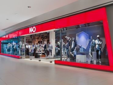Schaufenster mit großer roter Glasfassade, in der Schaufensterpuppen in Freizeitkleidung ausgestellt sind. Das Innere ist gut sichtbar und zeigt Kleiderständer. Das Logo des Ladens zeigt ein stilisiertes „K&Ö“.