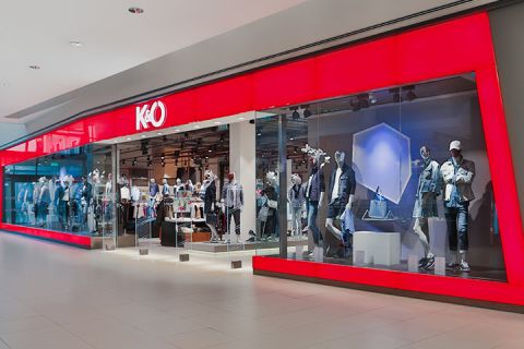 Schaufenster mit großer roter Glasfassade, in der Schaufensterpuppen in Freizeitkleidung ausgestellt sind. Das Innere ist gut sichtbar und zeigt Kleiderständer. Das Logo des Ladens zeigt ein stilisiertes „K&Ö“.