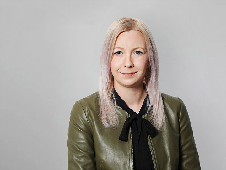 Porträt einer Frau mit glattem, hellblondem Haar, die eine grüne Lederjacke über einem schwarzen Oberteil trägt. Sie lächelt subtil vor einem schlichten, hellgrauen Hintergrund.