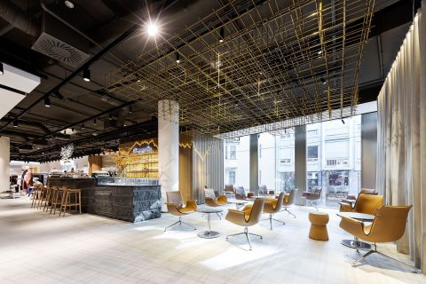 Moderner Loungebereich mit stilvollen braunen Stühlen und kleinen Tischen auf hellem Boden. Die Decke weist eine geometrische Goldstruktur auf. Große Fenster lassen viel Tageslicht herein und im Hintergrund ist eine Bar mit Hockern zu sehen.