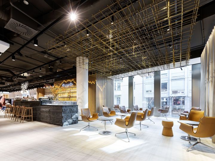 Moderner Loungebereich mit stilvollen braunen Stühlen und kleinen Tischen auf hellem Boden. Die Decke weist eine geometrische Goldstruktur auf. Große Fenster lassen viel Tageslicht herein und im Hintergrund ist eine Bar mit Hockern zu sehen.