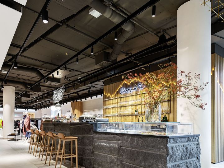 Modernes Café-Interieur mit Steintheke, Barhockern aus Holz und Zierpflanzen. Die Decke ist dunkel und mit Schienenbeleuchtung ausgestattet, im Hintergrund ist ein Sitzbereich mit Menschen zu sehen. Eine geometrische Deckeninstallation in Gold verleiht dem Raum eine stilvolle Note.