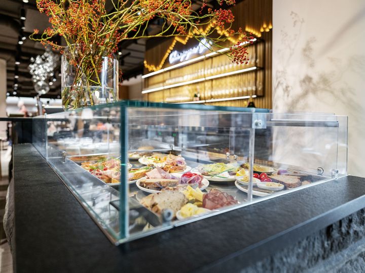 Glasvitrine in einem modernen Restaurant, in der verschiedene Speisen auf Tellern präsentiert werden, darunter Fleisch, Käse und Gemüse. Oben liegen dekorative Zweige mit roten Beeren. Holzwand mit beleuchteter Beschilderung im Hintergrund.
