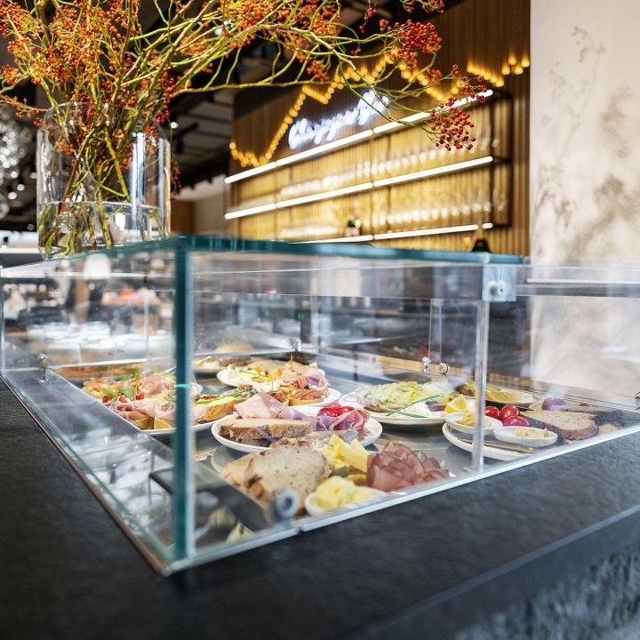 Glasvitrine in einem modernen Restaurant, in der verschiedene Speisen auf Tellern präsentiert werden, darunter Fleisch, Käse und Gemüse. Oben liegen dekorative Zweige mit roten Beeren. Holzwand mit beleuchteter Beschilderung im Hintergrund.