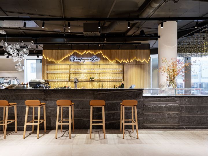 Eine moderne Champagnerbar mit einer eleganten Steintheke und fünf braunen Hockern. Hinter der Theke sind auf Regalen Gläser und Flaschen ausgestellt. Auf einem Neonschild steht „Champagne Bar“. Eine dekorative Leuchte und ein Blumenarrangement verleihen dem Raum Eleganz.