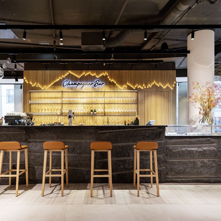 Eine moderne Champagnerbar mit einer eleganten Steintheke und fünf braunen Hockern. Hinter der Theke sind auf Regalen Gläser und Flaschen ausgestellt. Auf einem Neonschild steht „Champagne Bar“. Eine dekorative Leuchte und ein Blumenarrangement verleihen dem Raum Eleganz.