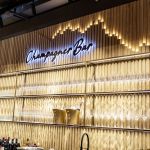 Champagnerbar mit Holzwandpaneelen und Regalen mit zahlreichen Champagnergläsern. Ein Neonschild darüber lautet „Champagne Bar“ und das gesamte Dekor ist mit goldenen Akzenten versehen.