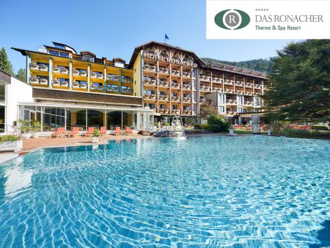Außenansicht des Das Ronacher Therme & Spa Resorts mit einem großen Swimmingpool mit Sonnenliegen drumherum. Das Resortgebäude hat mehrere Stockwerke mit gelben und braunen Fassaden und im Hintergrund sind Berge zu sehen.
