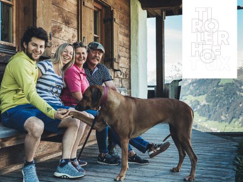 Vier Personen sitzen mit einem großen braunen Hund auf einer Holzveranda in den Bergen. Sie lächeln und halten den Hund an der Hand. Ein Logo in der oberen rechten Ecke lautet „TIROLERHOF TUX“ mit einer Berglandschaft im Hintergrund.
