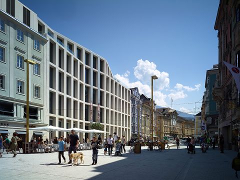 Eine belebte Stadtstraße mit modernen und traditionellen Gebäuden unter einem klaren blauen Himmel. Menschen gehen spazieren und sitzen in Straßencafés. Ein Mann geht mit einem Hund spazieren und im Hintergrund erheben sich Berge.
