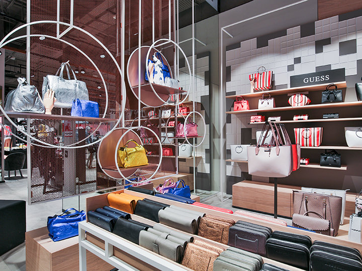 Eine stilvolle Verkaufsausstellung mit Handtaschen in verschiedenen Farben und Stilen verfügt über Regale und runde Hängevorrichtungen. Das helle, moderne Ladeninnere zeichnet sich durch ein auffälliges Guess-Schild zwischen den ordentlich angeordneten Taschen aus.