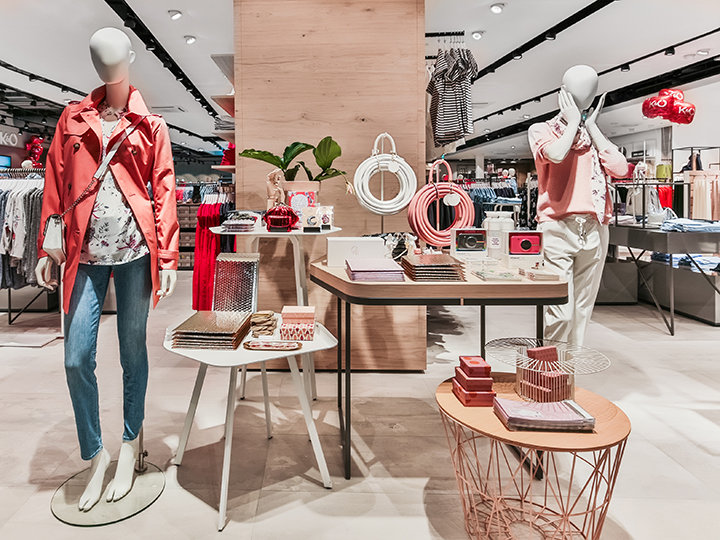 Ein stilvolles Schaufenster für ein Bekleidungsgeschäft mit Schaufensterpuppen in lässiger Frühlingskleidung. Auf Tischen werden Accessoires wie Handtaschen und Schmuck ausgestellt. Das Geschäft ist gut beleuchtet und gut organisiert, was eine moderne, einladende Einkaufsumgebung schafft.