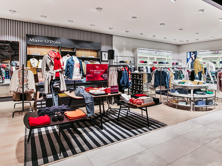 Ein modernes Bekleidungsgeschäft mit ordentlich angeordneten Regalen und Auslagen mit bunten Hemden, Jacken und Hosen. Das Geschäft ist hell beleuchtet und auf dem Boden liegt ein gestreifter Teppich. Im Hintergrund sind Markenschilder wie „Mare O'Polo“ zu sehen.