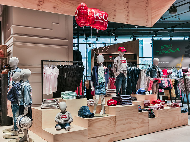 Schaufenster eines Kinderbekleidungsgeschäfts mit Schaufensterpuppen in legerer Kleidung, darunter Jeans und rote Hemden. Die Regale sind voll mit gefalteter Kleidung und Accessoires. Rote Luftballons hängen darüber und im Hintergrund steht auf einem Neonschild „STAY JUNGLE“.